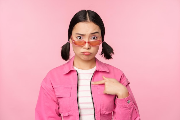 Portrait of asian girl looks confused and points at herself perplexed face stares with disbelief at camera stands over pink background