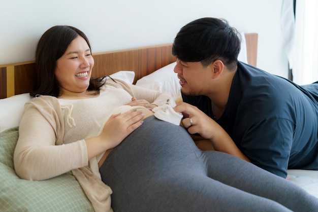 Portrait of Asian happy pregnant woman and of her husband holding baby shoes on bed