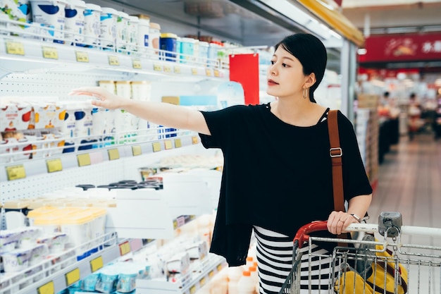 Portrait asian pregnant girl take yogurt at supermarket milk department. stylish motherhood woman with shopping cart for products from refrigerator in hyper market. Buying products at grocery store