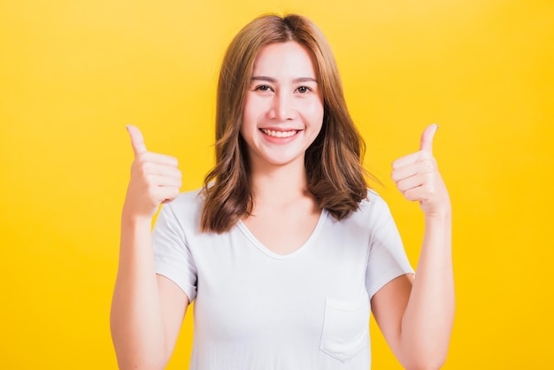 Portrait Asian Thai beautiful happy young woman smiling wear white t-shirt standing successful woman giving two thumbs up gesture sign in studio shot, isolated on yellow background with copy space