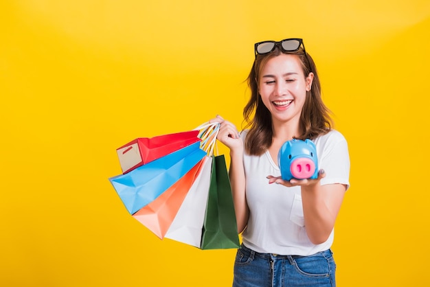 Portrait Asian Thai beautiful young woman happy hold colorful shopping bags and Piggybank Saving money, studio shot isolated on yellow background, with copy space