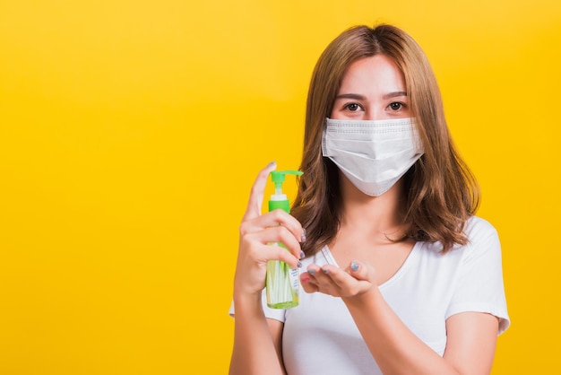 Portrait Asian Thai beautiful young woman wear mask protection clean corona virus on hand with alcohol gel or antibacterial soap sanitizer, studio shot isolated on yellow background, with copy space