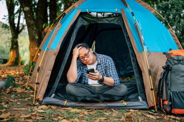 Portrait of Asian traveler man glasses using smartphone in tent camping Outdoor traveling camping and lifestyle concept