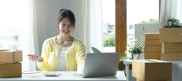 A portrait of Asian woman ecommerce employee sitting in the office full of packages on the table using a laptop and calculator for SME business ecommerce technology and delivery business