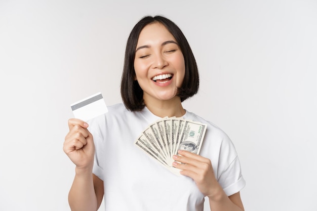 Portrait of asian woman smiling holding credit card and money cash dollars standing in tshirt over white background