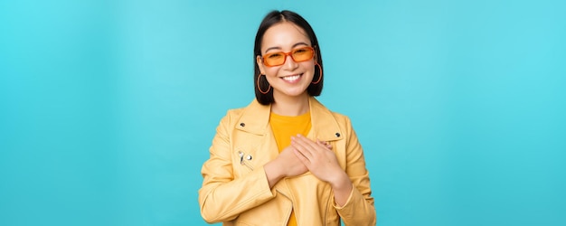 Portrait of asian woman smiling holding hands on heart and looking with tenderness care at camera thankful emotion standing over blue background