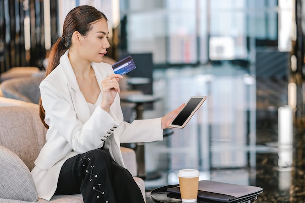 Portrait Asian woman using credit card with technology tablet for online shopping in modern lobby or working , coffee cup with computer laptop, technology money wallet and online payment 