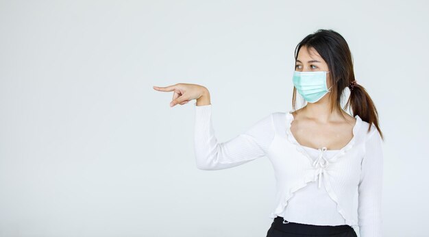 Portrait Asian woman wearing a face mask poses, pointing advertisement on white background, blank copy space with isolated white background cutout.