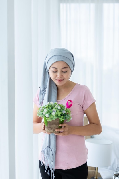 Portrait of a asian women disease mammary cancer patient Holding flower pots with pink ribbon wearing headscarf After treatment to chemotherapy at the window In the bedroom at the house