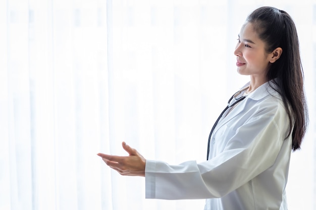 Portrait of asian young female doctor smiling face with stethoscope pointing with hand with copy space in hospital background.