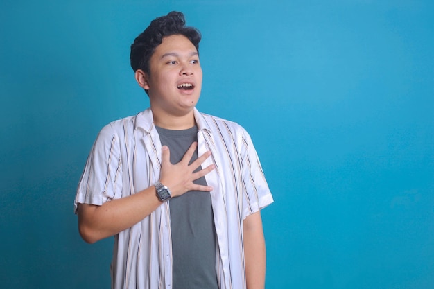 Portrait of Asian young man shocked and surprised while looking to copy space over blue background