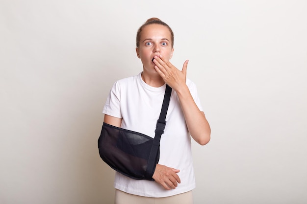 Portrait of astonished shocked woman with bun hairstyle wearing casual t shirt posing isolated over white background covering mouth with palm looks with big eyes being in sling for injured arm