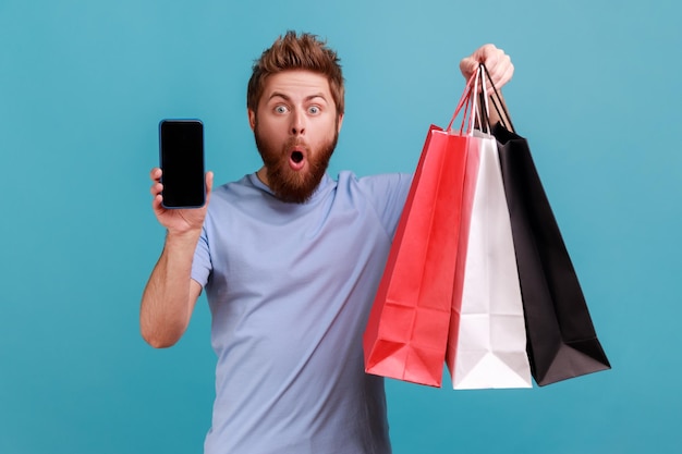 Portrait of astonished surprised bearded man standing with open mouth showing shopping bags and smart phone with blank screen for advertisement. Indoor studio shot isolated on blue background.