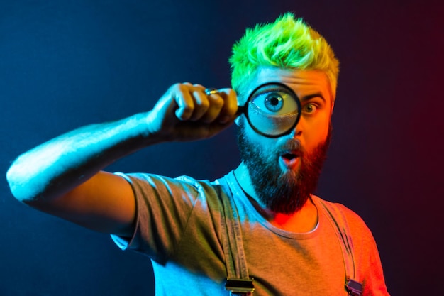 Portrait of astonished young adult man in denim overalls standing, holding magnifying glass and looking at camera with big zoom eye and surprised face. Colorful neon light, indoor studio shot.