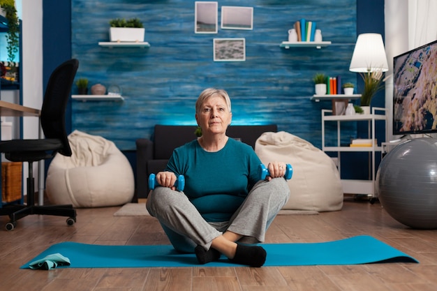 Portrait of athlete senior woman looking into camera sitting in lotus position on yoga mat in living room during wellness workout