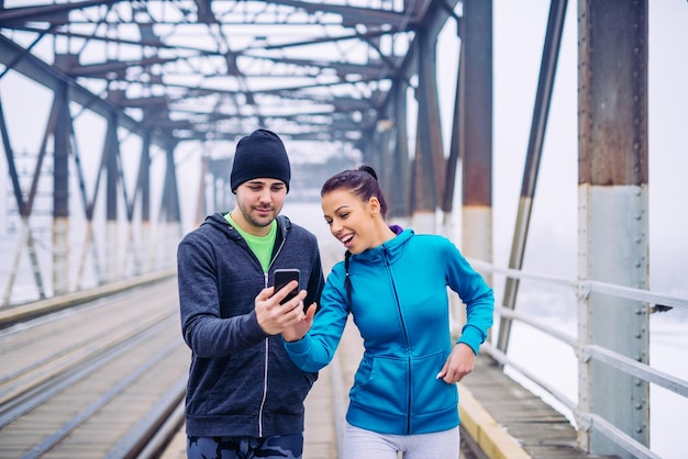 Portrait of an athletic couple having fun with mobile phone on the train bridge in the break