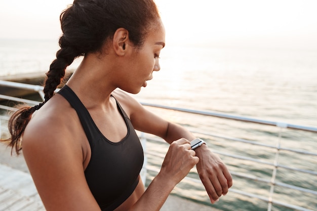 Portrait of athletic pretty woman in sportive clothes looking at her wristwatch while standing by seaside in morning