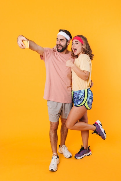 Portrait of athletic young couple wearing headbands smiling and taking selfie photo on cellphone isolated over yellow wall