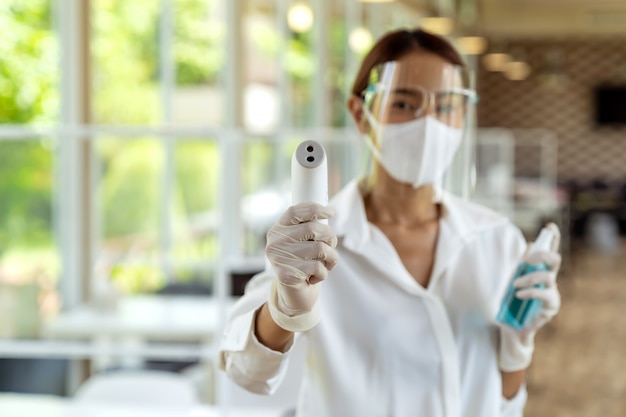 Portrait attractive asian waitress wear face mask and face shield hold thermometer and alcohol gel sanitizer for cleaning and hygiene with indoor restaurant background. New normal restaurant concept.