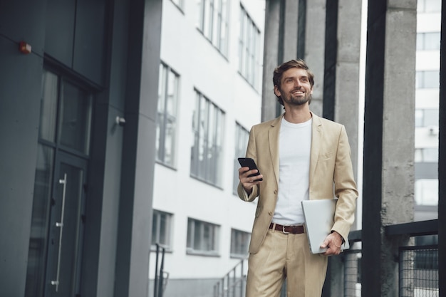 Portrait of attractive bearded man in stylish suit carrying laptop and smartphone on street