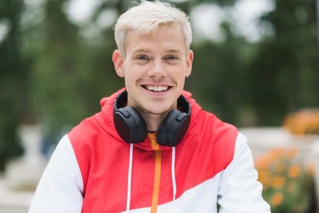 Portrait of attractive blond handsome man with healthy toothy smile with headphones on neck relaxing in the park of the city Street fashion style People lifestyle and technology concept