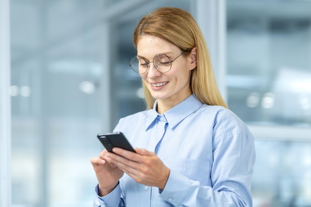 Portrait of attractive blonde woman in round eyeglasses looking down at cell phone in hands and