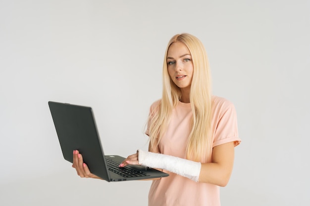 Portrait of attractive blonde young woman with broken arm wrapped in plaster bandage holding laptop and looking at camera standing on white background