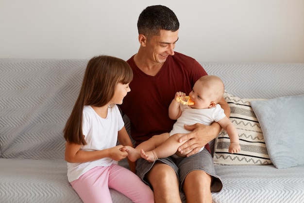 Portrait of attractive brunette man wearing maroon t shirt spending time with his children, family sitting on sofa, expressing positive emotions, enjoying weekend together.