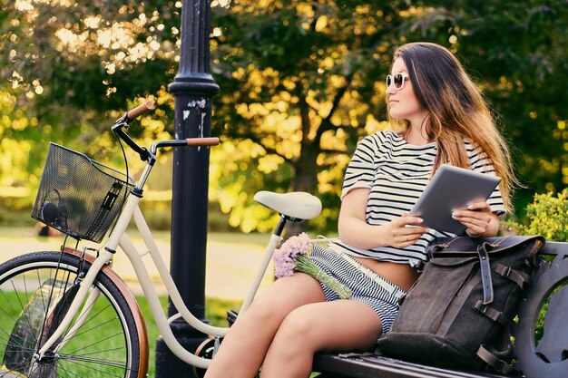 Portrait of an attractive brunette sits on a bench with a bicycle in a city park.