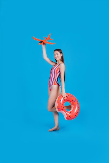 Portrait of attractive brunette woman in colored swimsuit holding rubber rings and parper plane posing sexy on camera Summer trendy beach style vacation at the resort Studio on a blue background