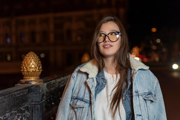 Portrait of attractive brunette woman in denim clothes evening city background Stylish girl on night city street