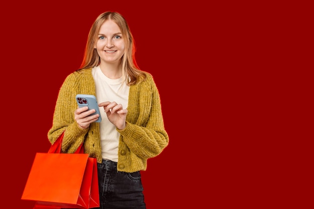 Portrait of attractive cheerful blonde woman using device holding bags order delivery isolated over red pastel color background