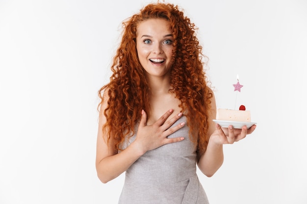 Portrait of an attractive cheerful young woman with long curly red hair standing isolated, celebrating birthday with a cake