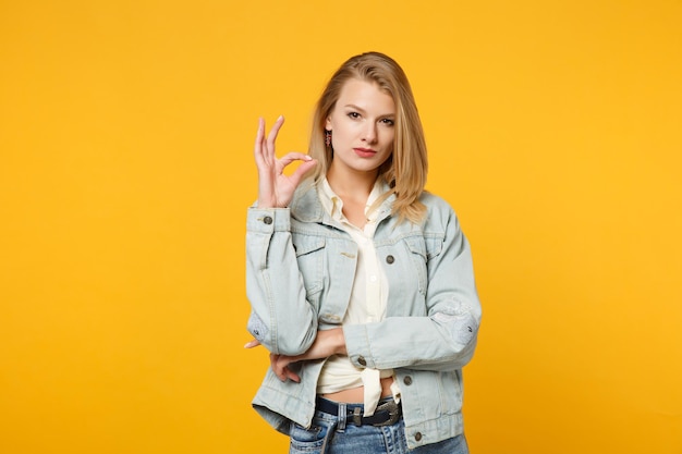 Portrait of attractive confident young woman in denim casual clothes looking camera, showing OK gesture isolated on yellow orange background in studio. People lifestyle concept. Mock up copy space.