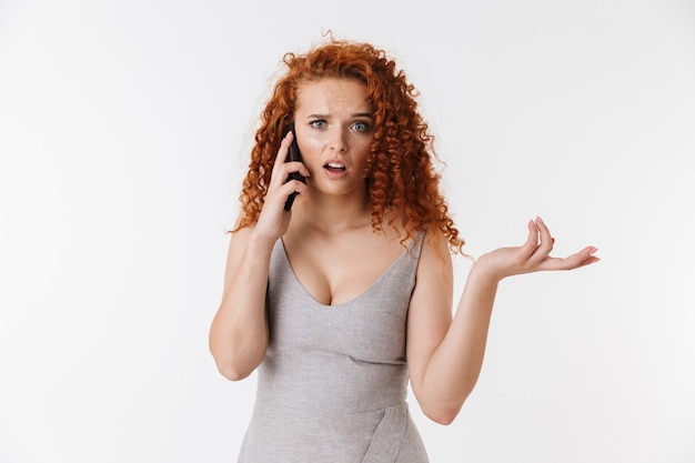 Portrait of an attractive confused young woman with long curly red hair standing isolated, using mobile phone