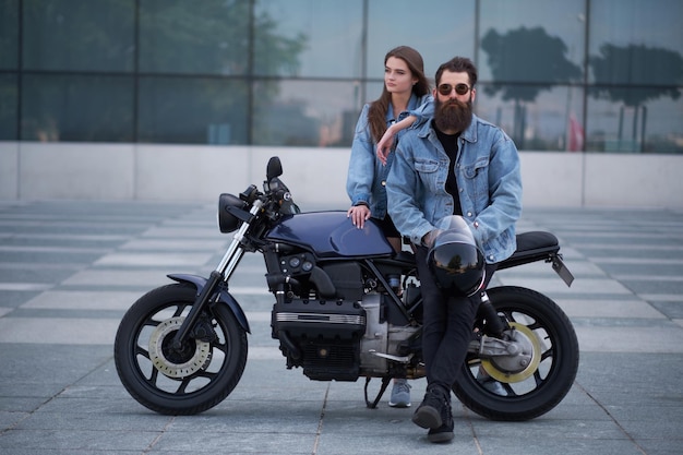 Portrait of attractive couple in denim jackets with motorbike near big glass building at city centre.