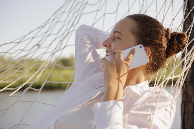 Portrait of attractive delighted woman wearing white shirt sitting in hammock on the bank of the river and talking smart phone with her friend looking away with joyful work