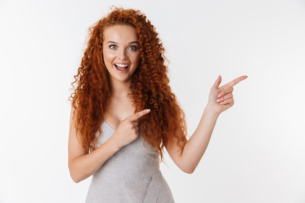 Photo portrait of an attractive excited young woman with long curly red hair standing isolated, presenting copy space, pointing