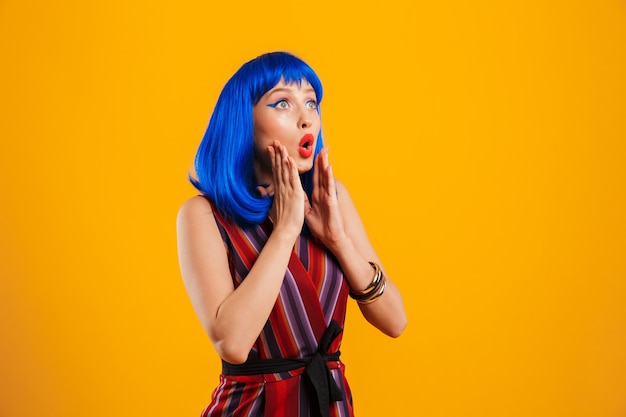 Portrait of an attractive funky young girl with blue hair wearing casual outfit standing isolated over yellow wall, looking away