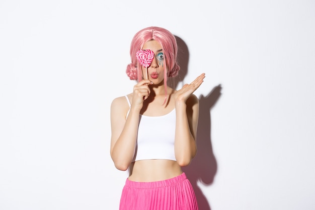 Portrait of attractive girl in pink wig holding cute candy over eye and pouting, wearing fairy costume on halloween, standing over white background.