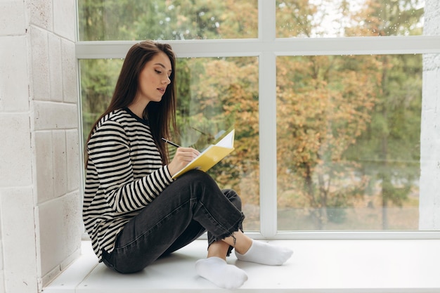 Portrait of an attractive girl sitting on the windowsill and doing homework in a notebook