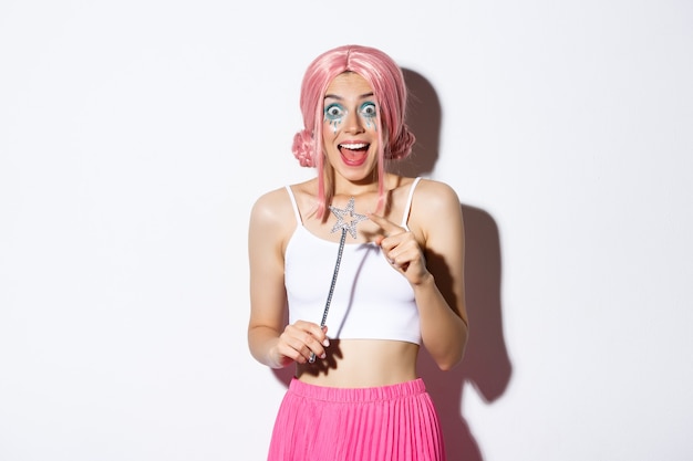 Portrait of attractive girl with pink wig and bright makeup, dressed up as a fairy for halloween party, holding magic wand and smiling