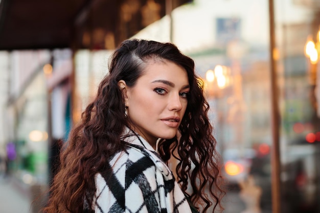 Portrait of attractive glamorous woman in elegant checkered coat in front of showcase lights, outdoors.