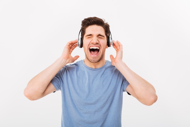 Portrait of attractive guy having short dark hair screaming his favorite song while enjoying music via earphones with closed eyes, isolated over white wall