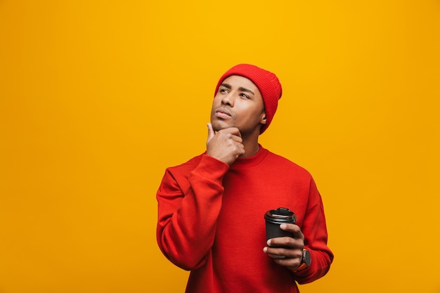 Portrait of an attractive pensive casual young african man standing over yellow wall, holding takeaway coffee cup