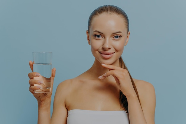 Portrait of attractive positive woman with glass of pure water standing against blue studio wall