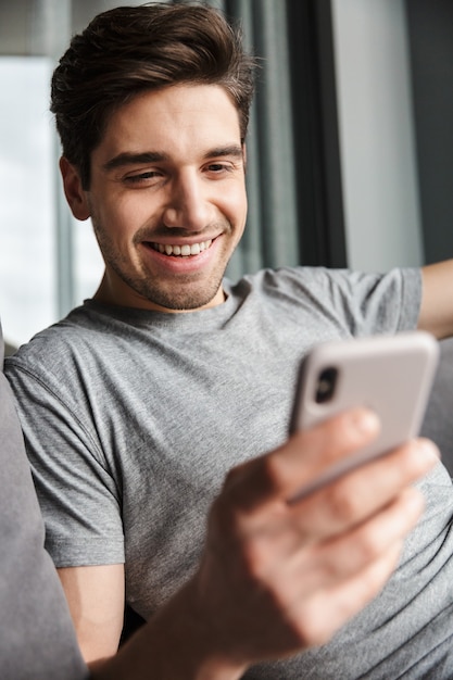 Portrait of an attractive smiling young bearded man wearing casual clothes sitting on a couch at the living room, using mobile phone