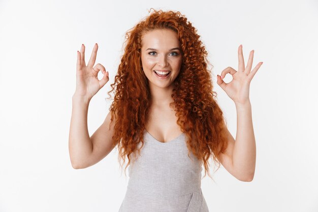 Portrait of an attractive smiling young woman with long curly red hair standing isolated, showing ok