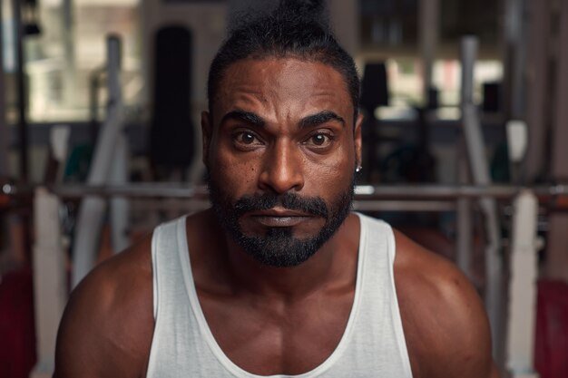 Portrait of an attractive sports man afro who stands in the gym in a white tshirt and looks directly...