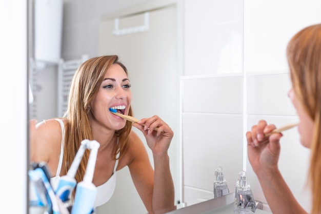 Portrait of attractive woman brushing teeth in bathroom and looking in the mirror at reflection. healthy teeth. Happy lovely young woman looking at the mirror and brushing teeth in bathroom